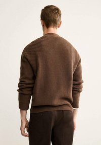 Maglione a coste marrone con maniche lunghe, dotato di collo rotondo e vestibilità comoda. Indossato con pantaloni marrone scuro.