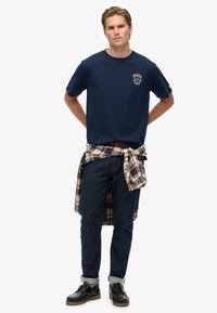 Joven de pie con las manos detrás de la espalda, vistiendo una camiseta navy blue de Superdry, jeans oscuros, zapatos negros y una camisa de cuadros atada alrededor de la cintura.