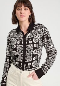 Chemise à motifs floraux noir et blanc avec col noir et fermeture à boutons sur le devant. Manches longues et design ajusté. Tissu léger.