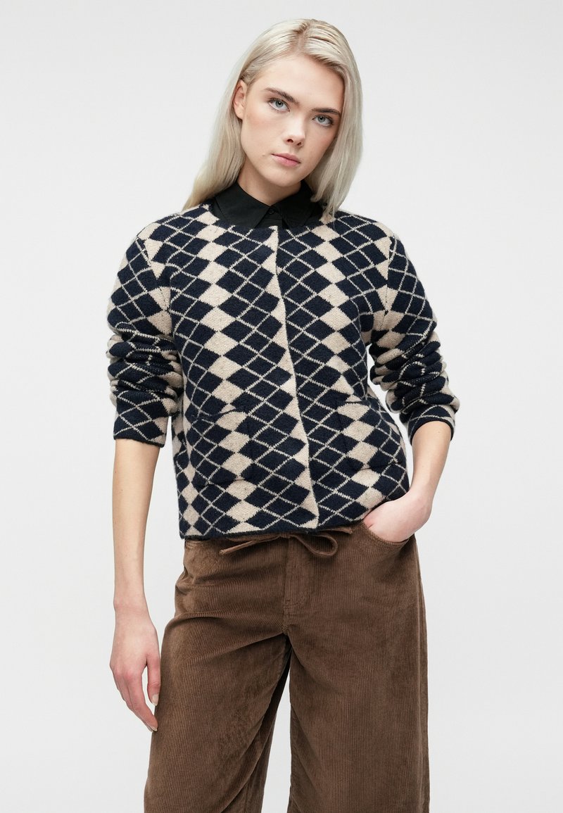 Jeune femme avec des cheveux blonds raides portant un pull au motif losange noir et beige et un pantalon en velours côtelé marron, avec une main dans la poche.