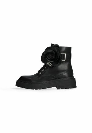 Botte de combat en cuir noir avec semelle épaisse, boucle argentée sur le côté, devant à lacets et grande fleur en tissu noir fixée près de la cheville.