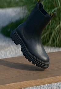 Musta nahkainen Chelsea-saapas paksulla urasoolilla, joka lepää puupinnalla, taustalla sumeita vihreitä kasveja.