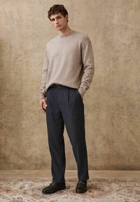 Pull en tricot beige texturé à col rond ; pantalon plissé navy ; mocassins noirs avec détails métalliques ; arrière-plan neutre.