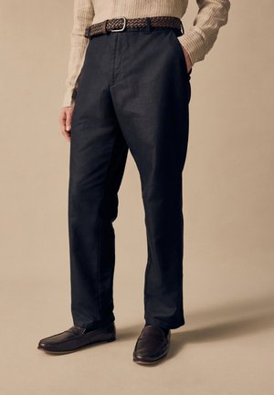 Hombre con pantalones negros, cinturón trenzado marrón, camisa beige de manga larga y mocasines marrón oscuro, de pie frente a un fondo beige liso.