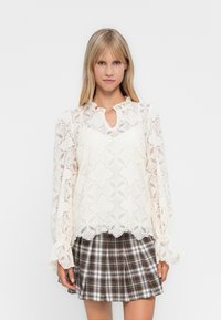 Blonde vrouw in een witte kanten blouse met lange mouwen en bloemenpatronen, met een bruin geruit plissé minirok, staand tegen een witte achtergrond.