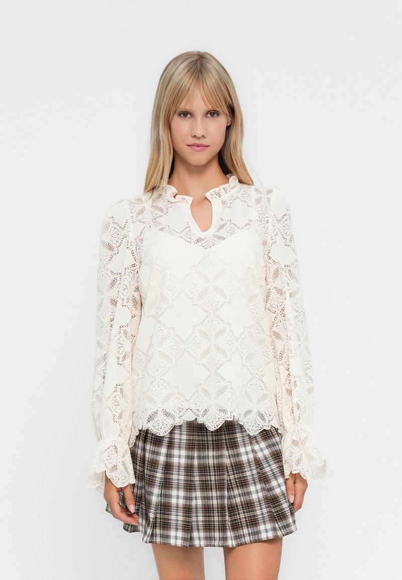 Blonde vrouw in een witte kanten blouse met lange mouwen en bloemenpatronen, met een bruin geruit plissé minirok, staand tegen een witte achtergrond.