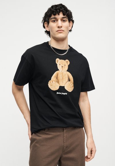 Junger Mann trägt ein schwarzes T-Shirt mit einer großen Grafik eines Teddybären und braune Hosen, steht vor einem schlichten weißen Hintergrund.