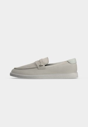 Lichtbeige suède loafer met een platte gestructureerde zool, een gevouwen riem aan de bovenkant en een klein metalen logo aan de zijkant.