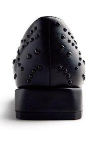 Chaussure noire en simili cuir avec un bout pointu, ornée de clous noirs brillants, présentant une semelle inférieure robuste et une finition mate.