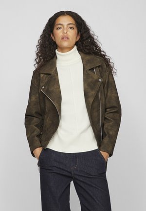 Chaqueta de motorista de imitación cuero marrón con un acabado ligeramente texturizado, detalles de cremallera y solapas anchas, llevada sobre un suéter de cuello alto blanco.