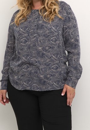 Woman wearing a long-sleeve navy blue blouse with white abstract circular patterns and black pants, standing against a plain white background.