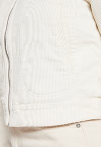 White corduroy jacket with a zip closure, ribbed texture, side pockets, and clean seam lines, paired with matching white fabric below.