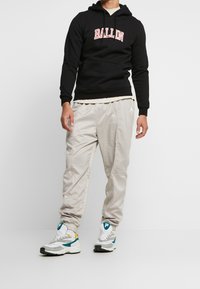 Schwarzer Hoodie mit "BALLIN" Grafik auf der Brust, kombiniert mit hellbeigen Jogginghosen. Die Schuhe sind in einer Mischung aus Weiß, Grau und Türkis gehalten.