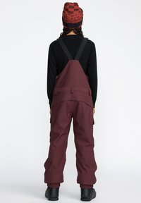 Burgundy snöoverall med justerbara svarta remmar, cargofickor och ribbade ärmslut. Kombinerad med en mönstrad röd och svart mössa.
