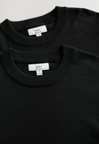 Deux pulls noirs à col rond étiquetés "Next", taille moyenne, coupe régulière, fabriqués au Bangladesh, posés à plat sur un fond clair.