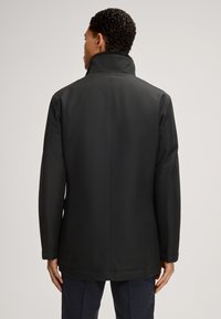 Schwarzer Mantel mit hohem Kragen, aus einem glatten, matten Material gefertigt. Verfügt über einen taillierten Schnitt und ein minimalistisches Design, geeignet für formelle Anlässe.