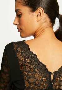 Top nero con dettagli in pizzo intricati, scollatura a forma di V e un bottone decorativo sul retro. Tessuto testurizzato con materiali a contrasto.
