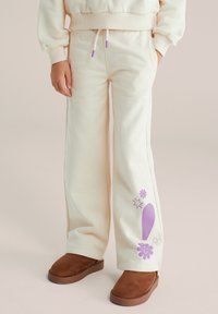 Kremaste trenirka z širokimi hlačnicami, elastičnim pasom, vijoličnim vezalko in igrivimi vijoličnimi grafičnimi motivi; kombinirane z rjavimi semiš natikači.