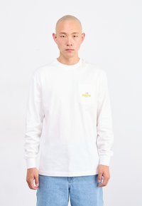 Un jeune homme à la tête rasée porte une chemise blanche à manches longues avec un logo sur la poche et un jean bleu clair, se tenant devant un fond blanc uni.
