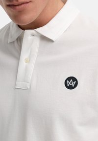 Hvit poloskjorte i pustende stoff med en strukturert overflate, med en svart brodert logo på venstre bryst og krage med to knapper.