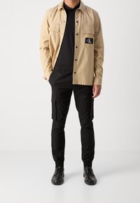 Beige Hemd mit Knopfleiste und zwei Brusttaschen, schwarzes Polo darunter, schwarze Cargohosen mit elastischen Bündchen und schwarze Schuhe.