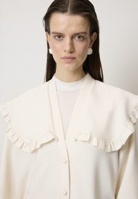 Chaqueta blanca con cuello rizado y botones delanteros, confeccionada en tejido suave. Llevada sobre una blusa ligera y texturizada.