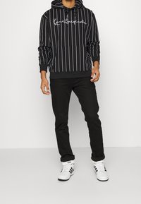 Mies, joka käyttää mustaa raitapaitaa valkoisella kirjotuksella, mustia housuja ja valkoisia Adidas-tennareita tasaisella valkoisella taustalla.