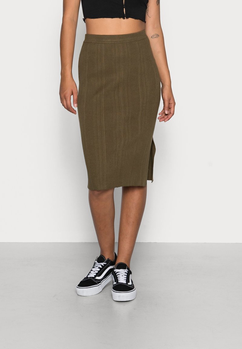 Personne portant une jupe midi côtelée olive avec une fente latérale, un haut court noir et des baskets lacées noires et blanches, debout sur un sol gris.