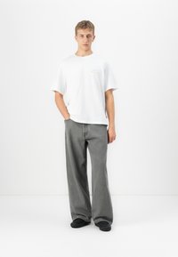 Ung mann står med den ene hånden i lommen, iført hvit oversized t-skjorte, løse grå jeans og svarte sko mot en ensfarget hvit bakgrunn.