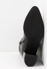 Botte en cuir noir avec un talon carré et épais, une fermeture éclair sur le côté et une semelle texturée, inclinée contre un coin blanc en arrière-plan.