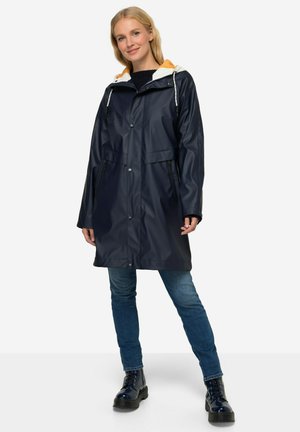 Navy waterproof coat with a zippered front, hood lined in orange, and two front pockets. Worn over blue jeans and black boots.