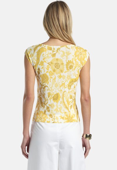 Frau mit langen blonden Haaren, die ein gelbes, floral gemustertes Top mit Kapuzenärmeln und eine weiße Hose trägt, von hinten gesehen.