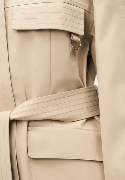 Κλικ σε κοντινό πλάνο λεπτομερειών μπεζ καπαρντίνας που δείχνει τη ζώνη, την τσέπη με καπάκι, τις ραμμένες ραφές και το μεταλλικό D-ring σε υφασμάτινη θηλιά.