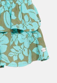 Υφασμα με στρώσεις με λουλουδένιο μοτίβο σε τιρκουάζ και ελιά, με ένα μικρό στρογγυλό μεταλλικό ταμπελάκι προσαρτημένο κοντά στο στρίφωμα.