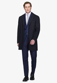 Mørk overfrakke over en marineblå stribet jakkesæt, hvid skjorte og sølv slips. Sorte lædersko fuldender det skræddersyede look.