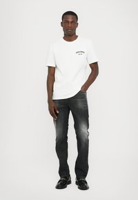 Camiseta blanca de manga corta con logo, combinada con jeans de mezclilla oscura que tienen un lavado desvaído y sutiles detalles desgastados. Zapatos negros.