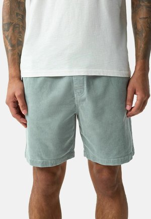 Homme portant un short en velours côtelé bleu clair et un t-shirt blanc, avec les bras tatoués visibles des coudes vers le bas.