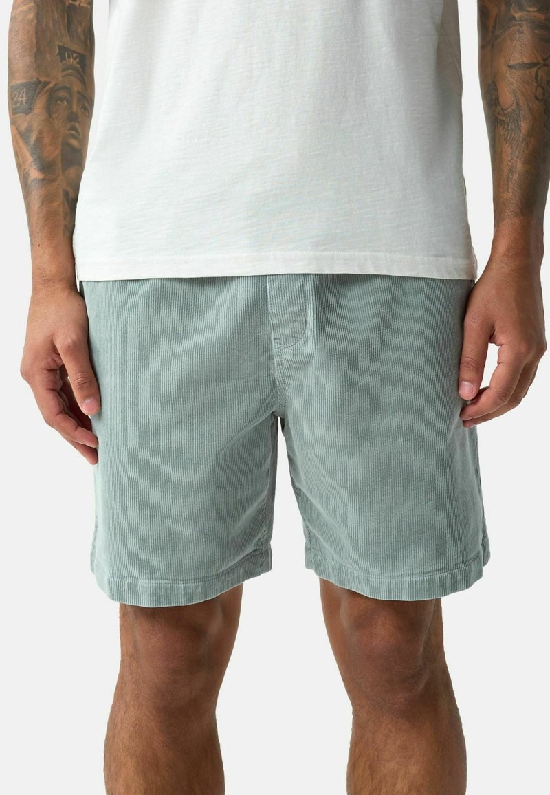 Homme portant un short en velours côtelé bleu clair et un t-shirt blanc, avec les bras tatoués visibles des coudes vers le bas.