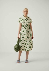 Donna che indossa un abito midi bianco con motivi a diamante verdi, tiene in mano una borsa trapuntata verde e indossa sandali aperti blu.