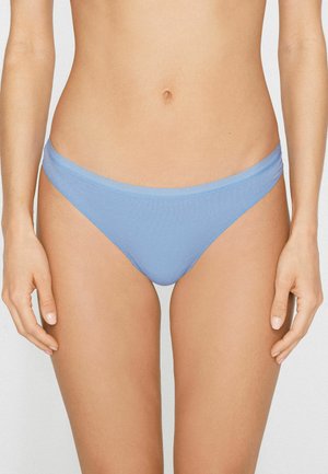 Kobieta w jasno-niebieskiej bieliźnie bikini stojąca na jednolitym tle, widoczna od talii do górnej części ud.