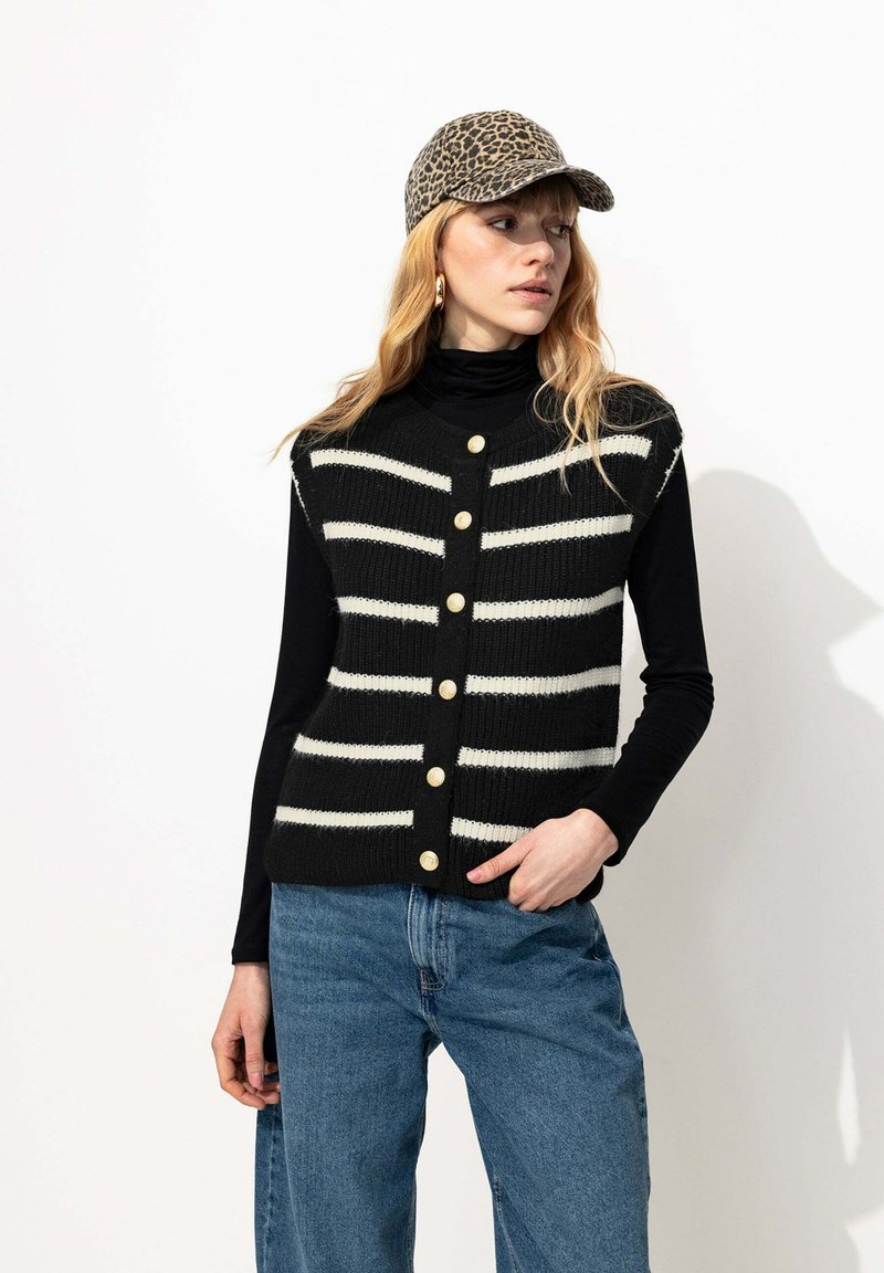 Gilet en tricot à rayures noires et blanches avec des boutons dorés, superposé sur un col roulé noir, associé à un jean en denim bleu et une casquette à imprimé léopard.