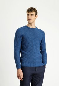 Mężczyzna w niebieskim, teksturowanym swetrze dzianinowym i ciemnych spodniach, stojący z jedną ręką w kieszeni, patrzący na bok na tle jednolitego tła.