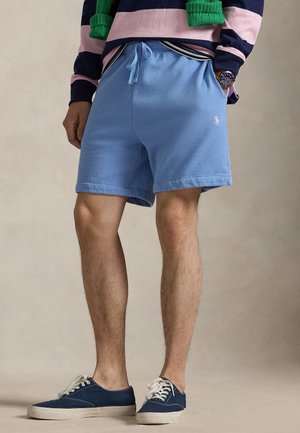 Uomo che indossa pantaloncini azzurri con coulisse, sneakers blu navy, un maglione a righe rosa e blu navy e un maglione verde drappeggiato sulle spalle.