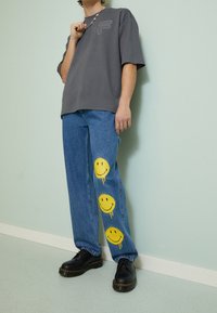Camiseta gris de gran tamaño con un pequeño logo, combinada con jeans de mezclilla azules que presentan gráficos de caras sonrientes amarillas en las piernas inferiores. Zapatos negros.