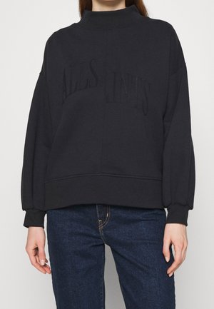 Femme portant un sweat-shirt noir oversize avec un texte en relief et un jean bleu sur un fond uni.
