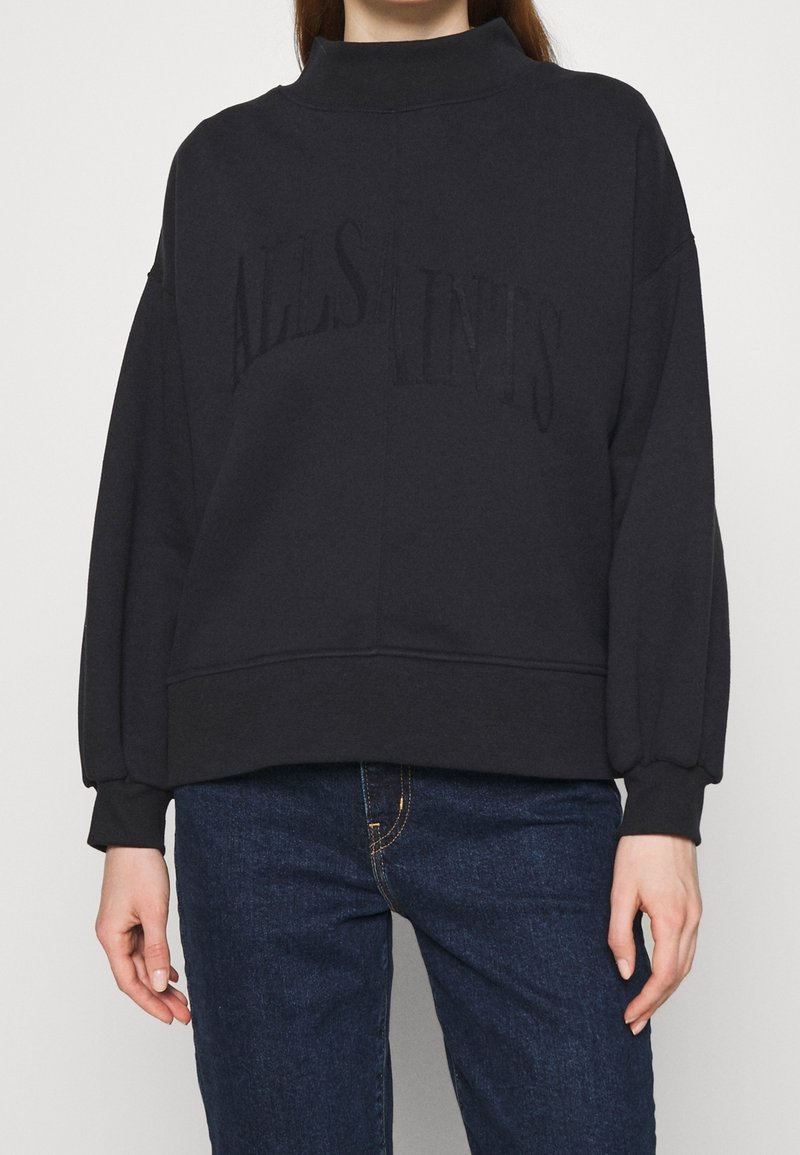 Femme portant un sweat-shirt noir oversize avec un texte en relief et un jean bleu sur un fond uni.