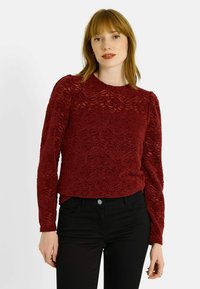 Haut en dentelle bordeaux à manches longues avec motif floral, encolure ronde, texture travaillée et coupe ajustée, porté avec un pantalon noir.