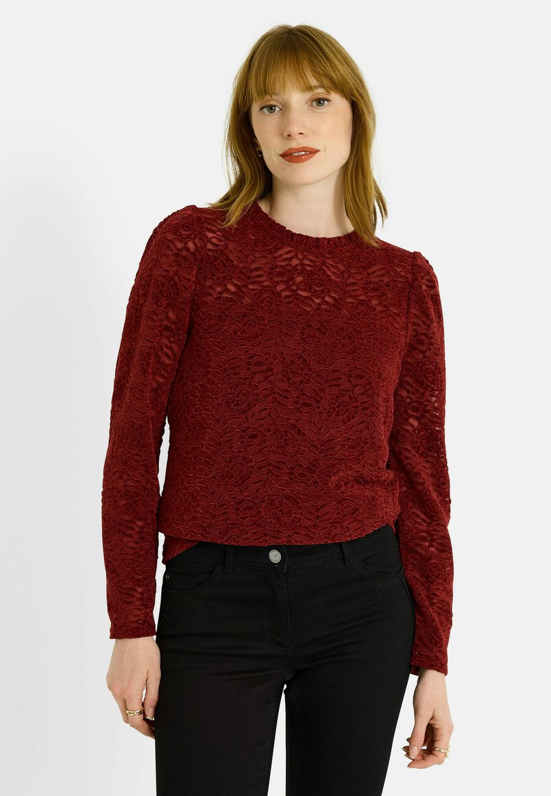 Haut en dentelle bordeaux à manches longues avec motif floral, encolure ronde, texture travaillée et coupe ajustée, porté avec un pantalon noir.