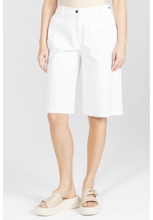 Osoba ubrana w białe szorty do kolan i beżowe klapki na platformie, stojąca na tle jednolitego jasnoszarego tła.