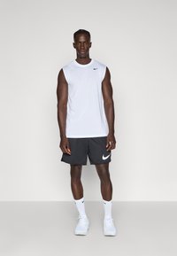Biele bezrukávové športové tričko s čiernym logom Nike; šedé športové šortky s bielym logom Nike; biele topánky; jednoduché pozadie.